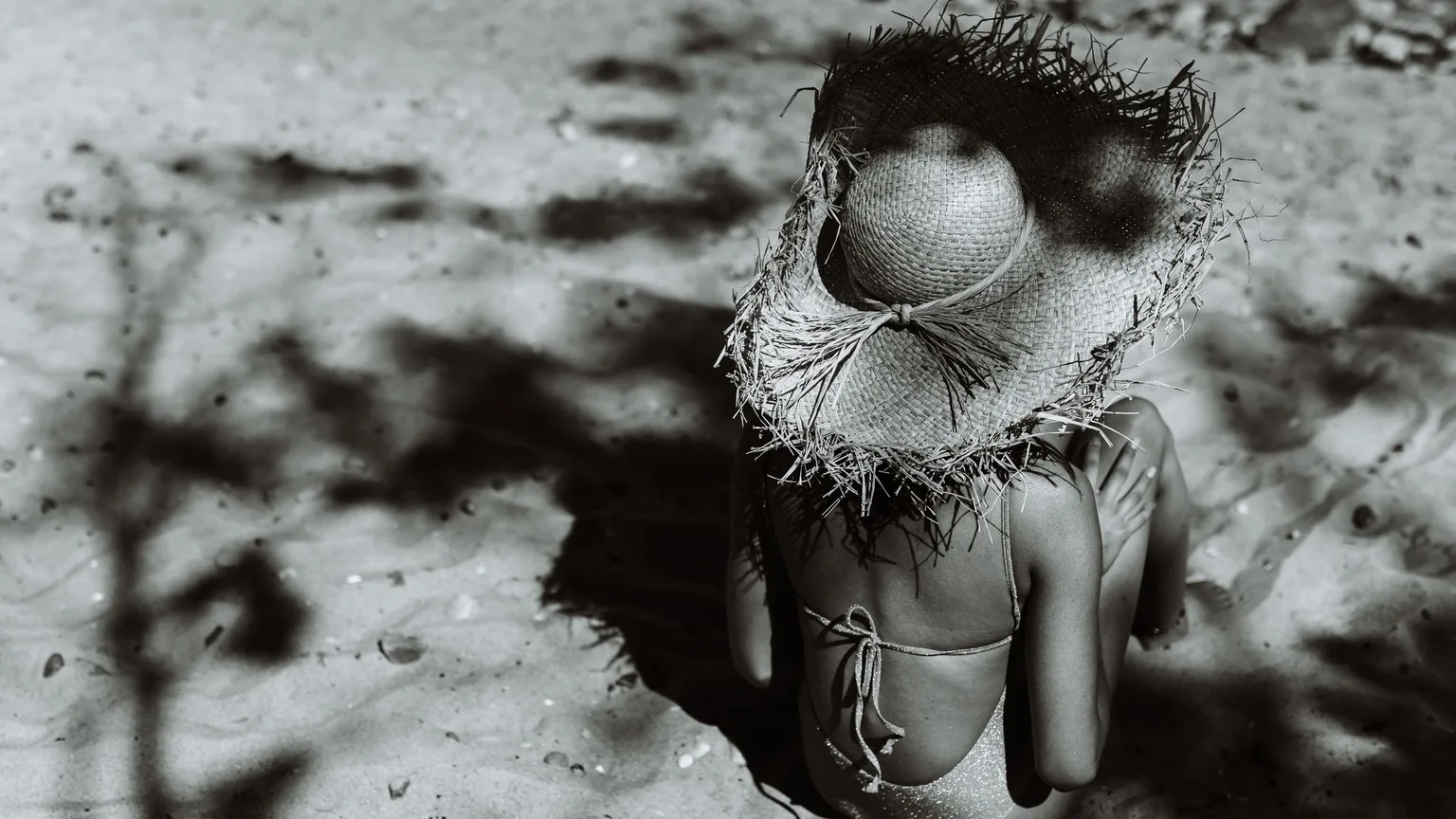 Overhead black and white photograph of a woman in a straw sun hat seated on sand with palm shadow